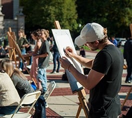 A man stands at an easel drawing outside on the Plaza of Low Library, joined by more than a dozen others also drawing at easels.